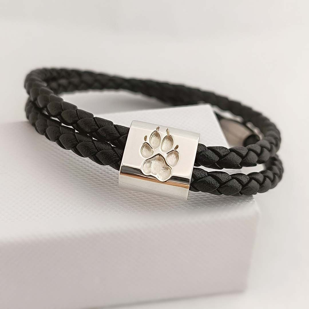 Schwarzes, geflochtenes Lederarmband mit silbernem Verschluss und eingravierter Pfote, auf weißem Untergrund