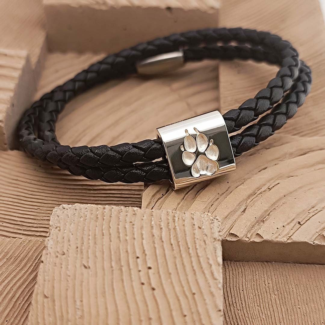 Schwarzes, doppelt gewickeltes Lederarmband mit silbernem Verschluss und eingravierter Pfote, auf Holzuntergrund