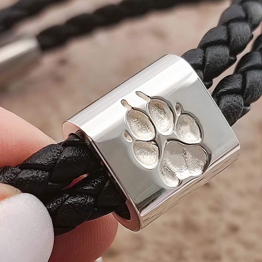 Nahaufnahme eines silbernen Verschlusses mit eingravierter Pfote auf schwarzem, geflochtenem Lederarmband