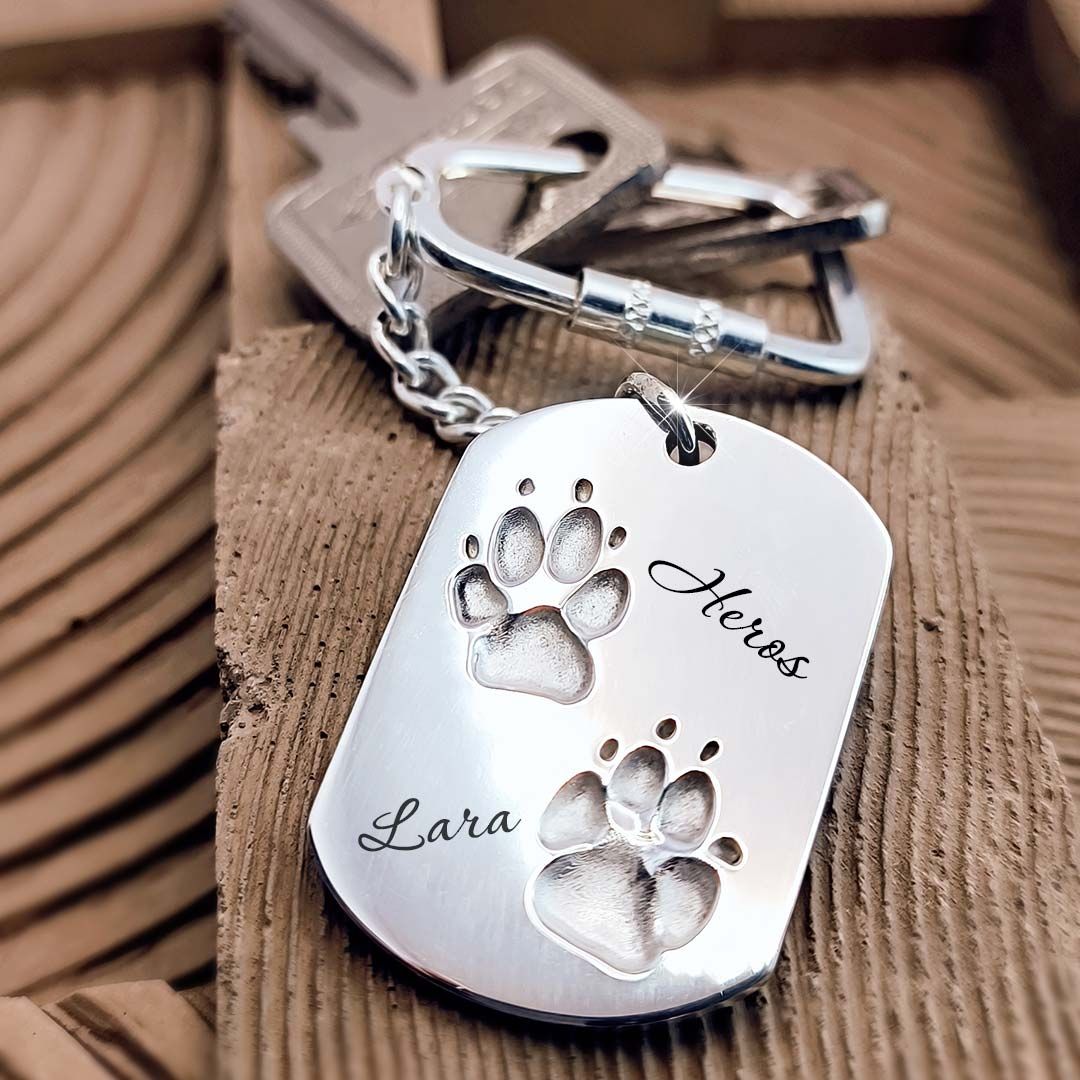 Silberner Schlüsselanhänger mit Gravuren Lara und Heros neben zwei Pfotenabdrücken, auf Holzuntergrund mit Schlüsseln im Hintergrund