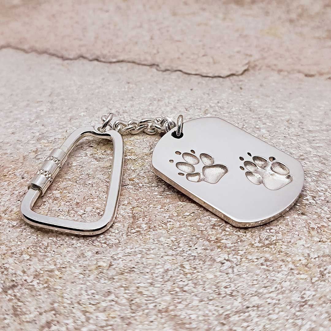 Silberner Schlüsselanhänger mit rechteckigem Anhänger und zwei eingravierten Pfotenabdrücken, auf Steinuntergrund