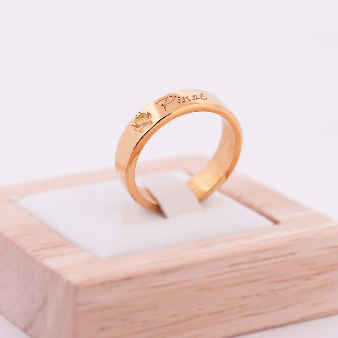 Goldener Ring mit Gravur Pinot und kleiner Pfote, präsentiert auf einem Holzringhalter mit weißem Einsatz