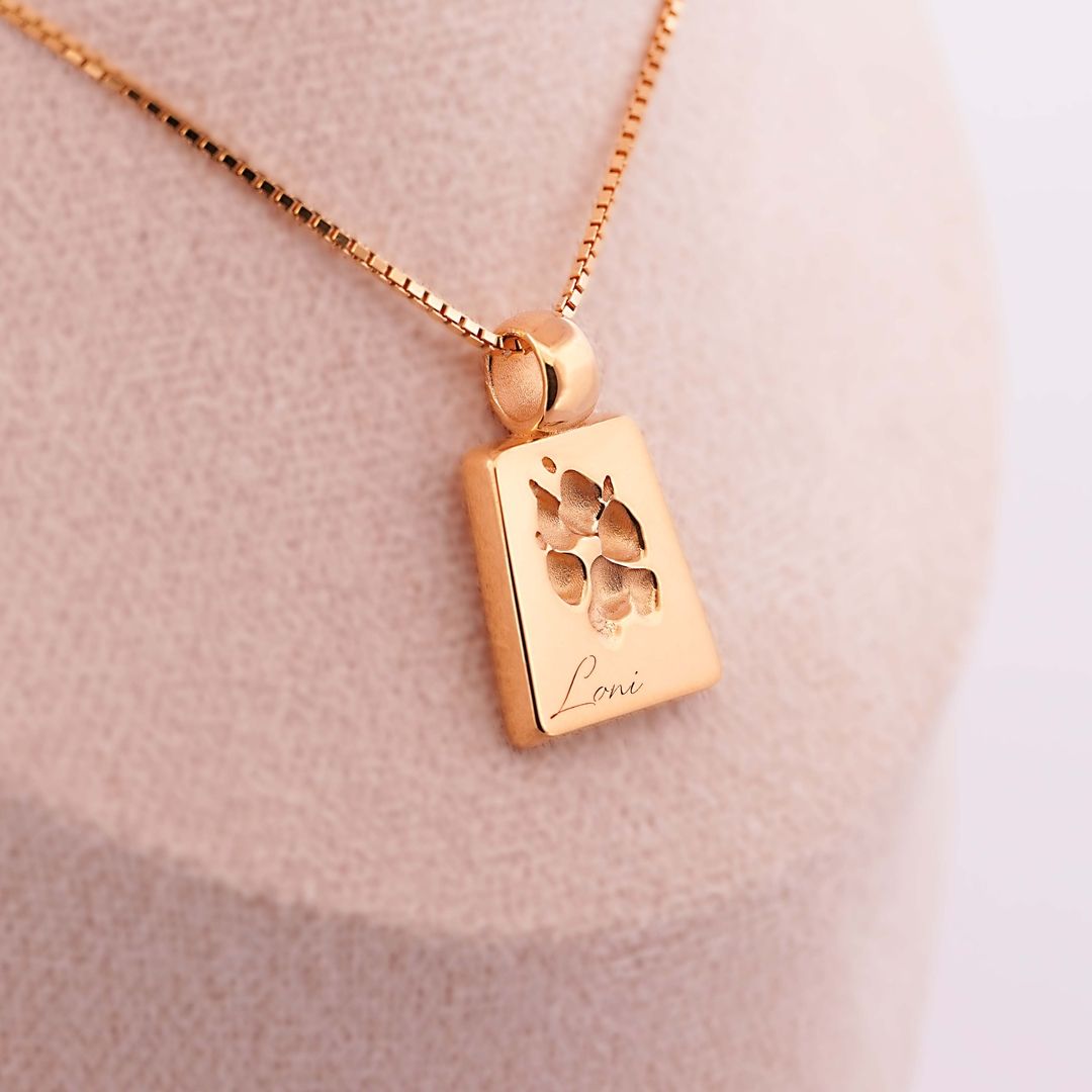 Rechteckiger, goldener Anhänger mit ausgeschnittener Pfotenform und Gravur Lani, an einer feinen Kette auf beigem Schmuckdisplay