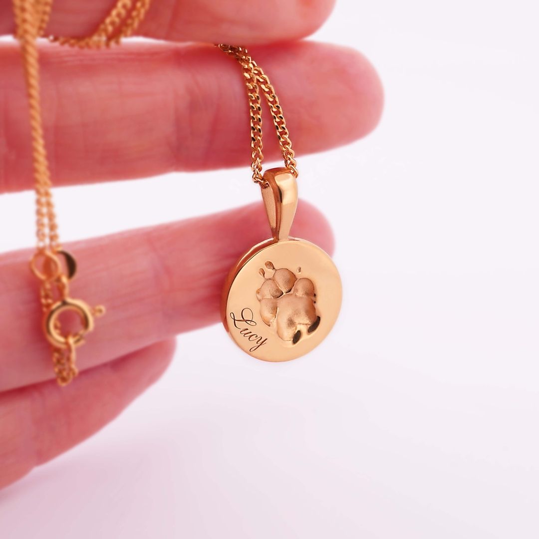 Runde, goldene Halskette mit eingravierter Pfotenform und Gravur Lucy, zwischen Fingern gehalten