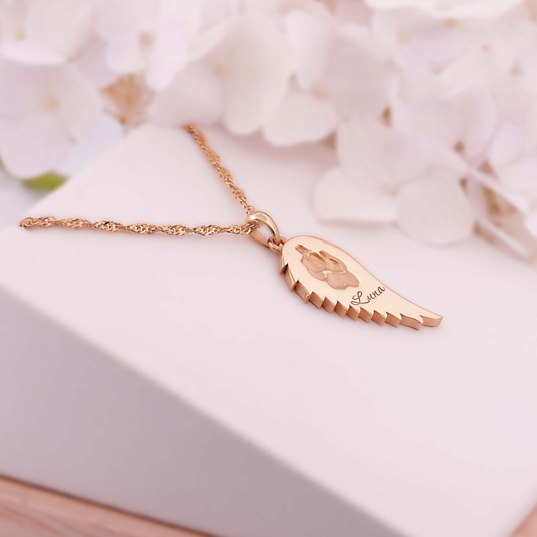 Goldene Halskette mit Flügelanhänger und Gravur Luna neben eingravierter Pfote, auf heller Schmuckbox mit Blüten im Hintergrund