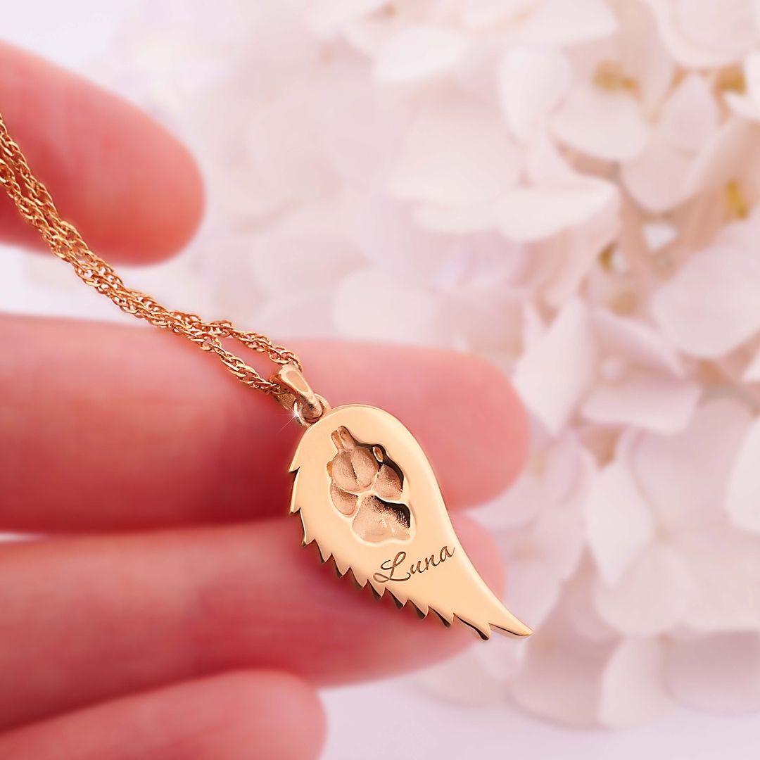 Goldene Halskette mit flügelähnlichem Anhänger, eingravierte Pfote und Name Luna, gehalten vor weißen Blüten