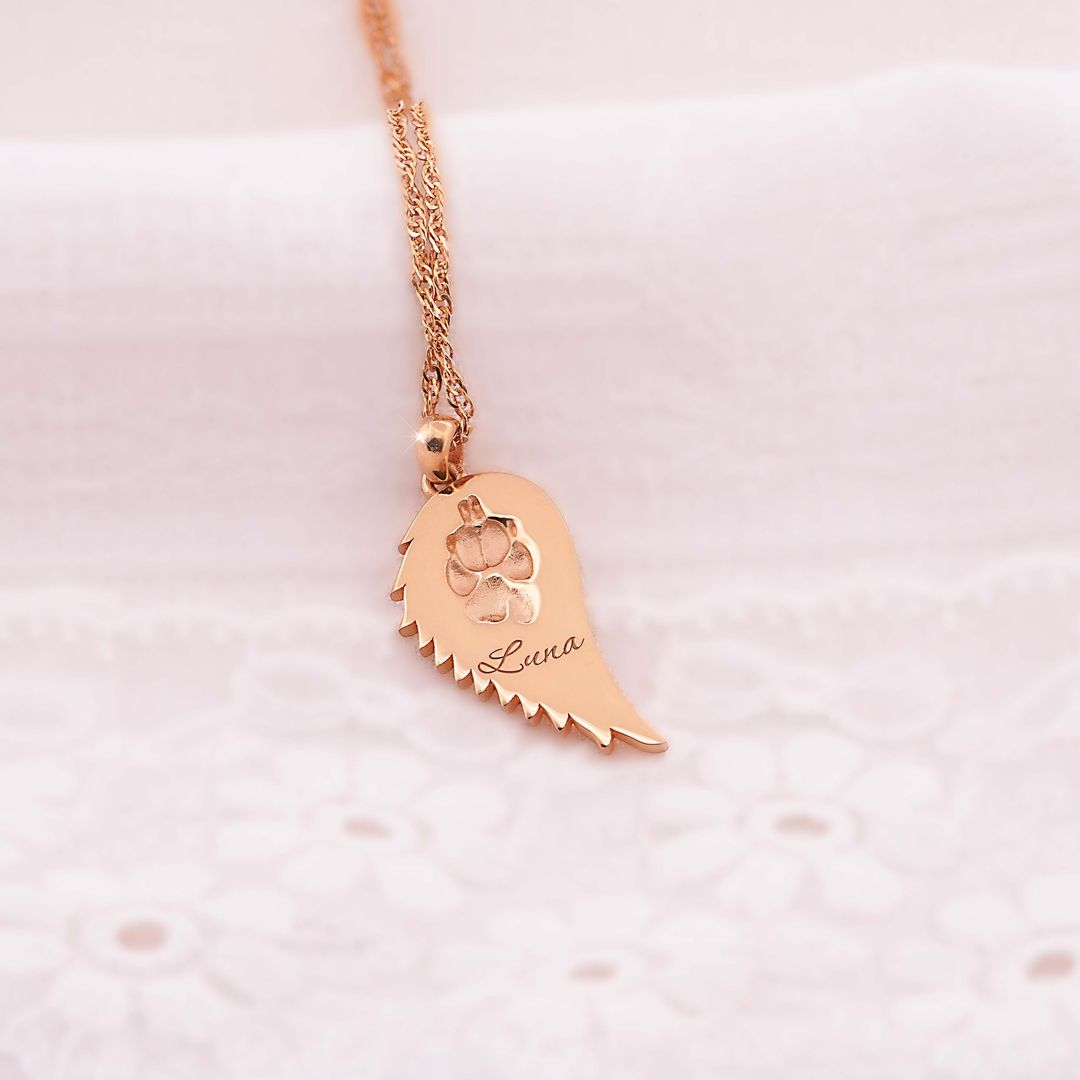Goldene Halskette mit Flügelanhänger, eingravierte Pfote und Name Luna, auf zarter weißer Spitze arrangiert