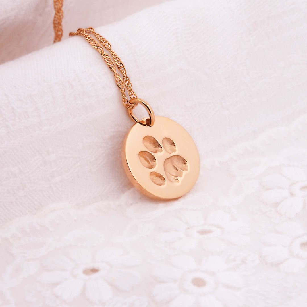 Goldene Halskette mit rundem Anhänger, der eine eingravierte Pfote zeigt, auf weißer Spitze drapiert