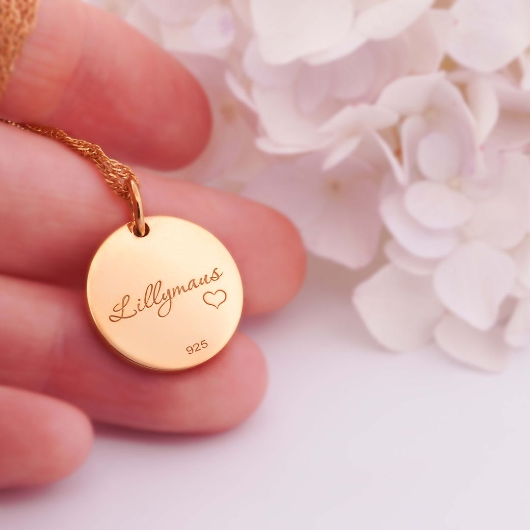 Runde, goldene Halskette mit Gravur Lillymaus und kleinem Herzsymbol, gehalten vor weißen Blüten